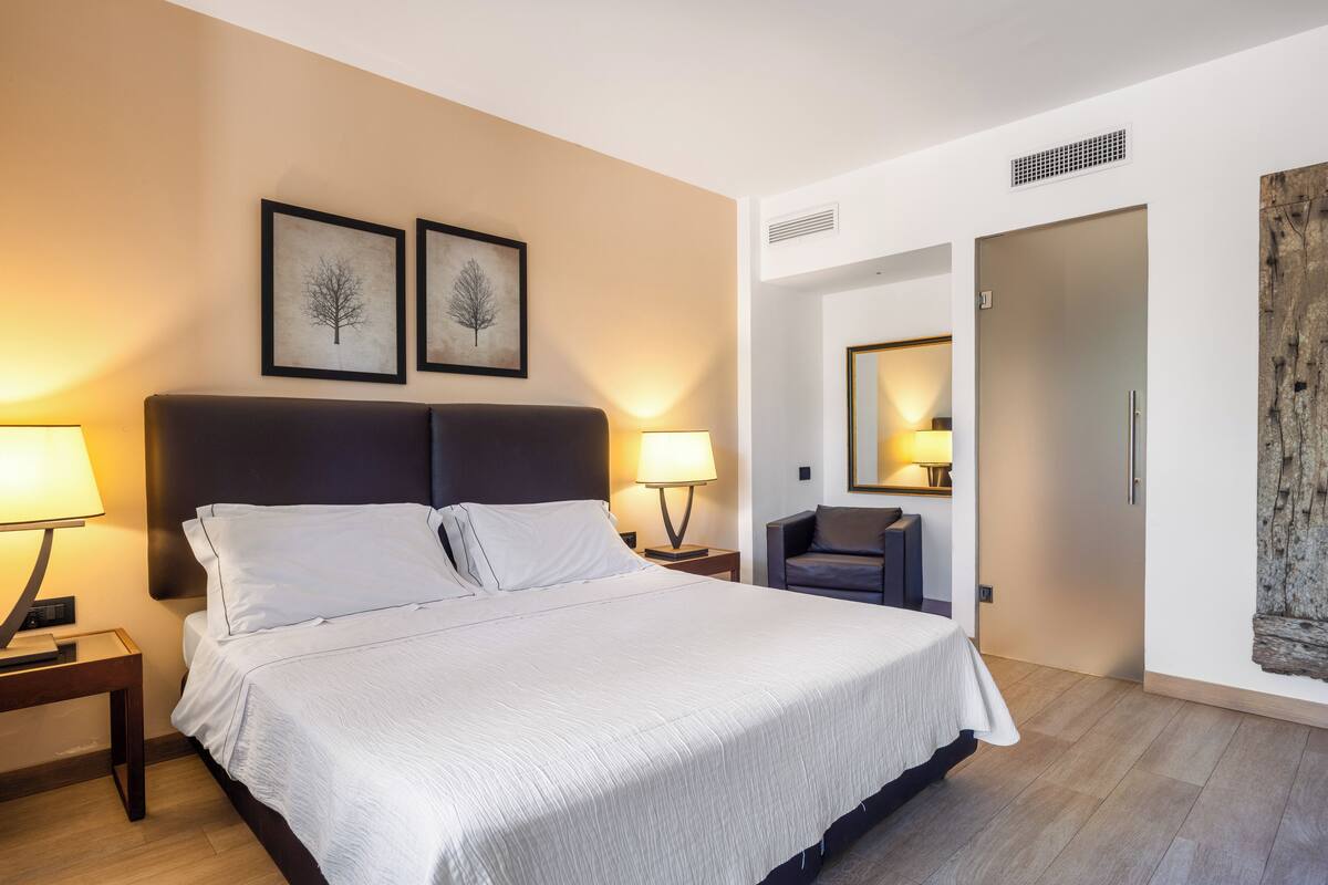 A neatly arranged hotel room with a large bed, two bedside lamps, a chair, and a door leading to another room.
