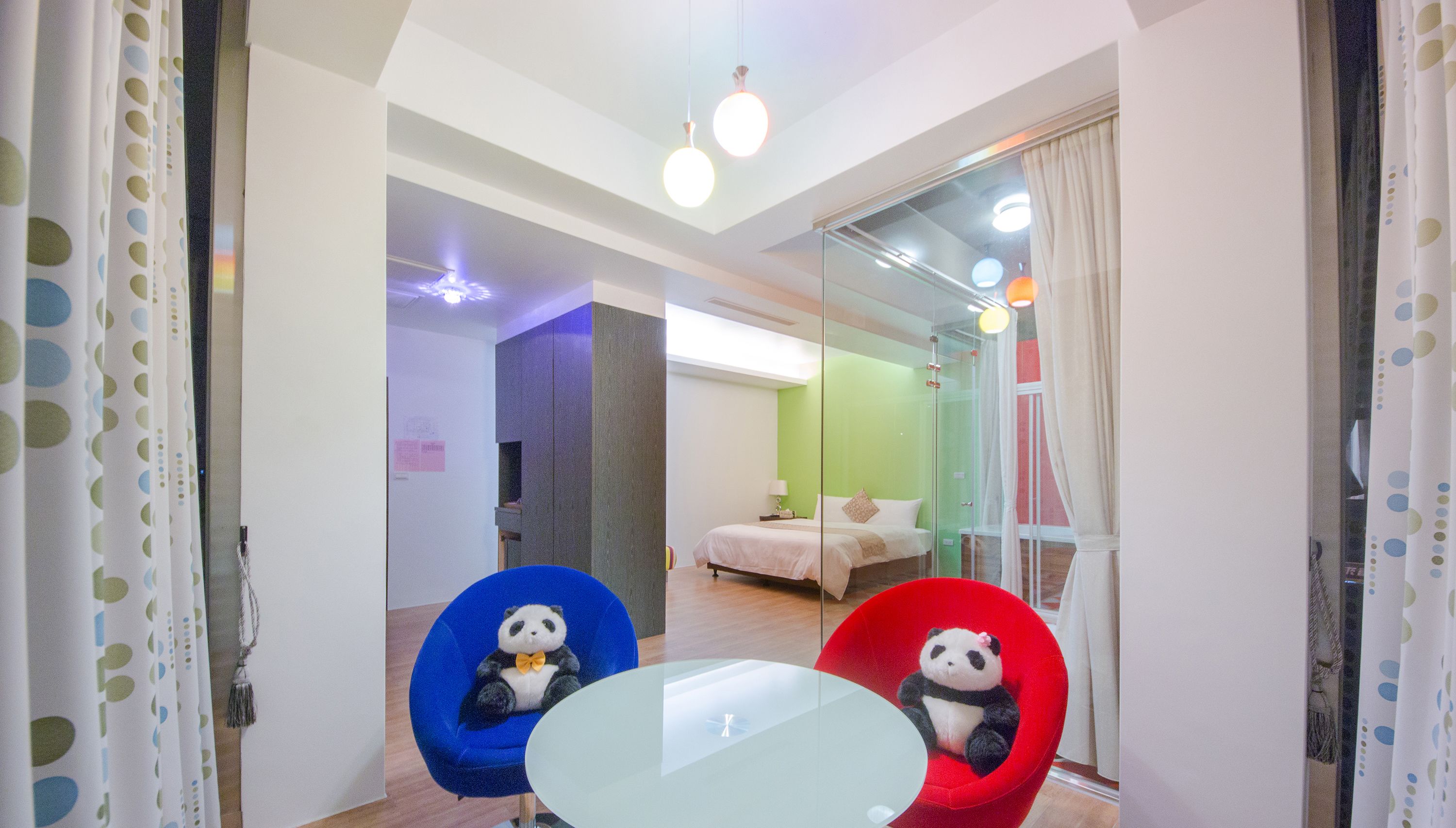 Habitación de hotel con mampara de vidrio, una cama, una mesita pequeña y dos sillas con cojines con temática de panda.