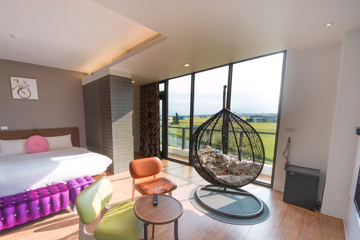 A modern hotel room with a large bed, a hanging chair, a small table, and a view of the outdoors.
