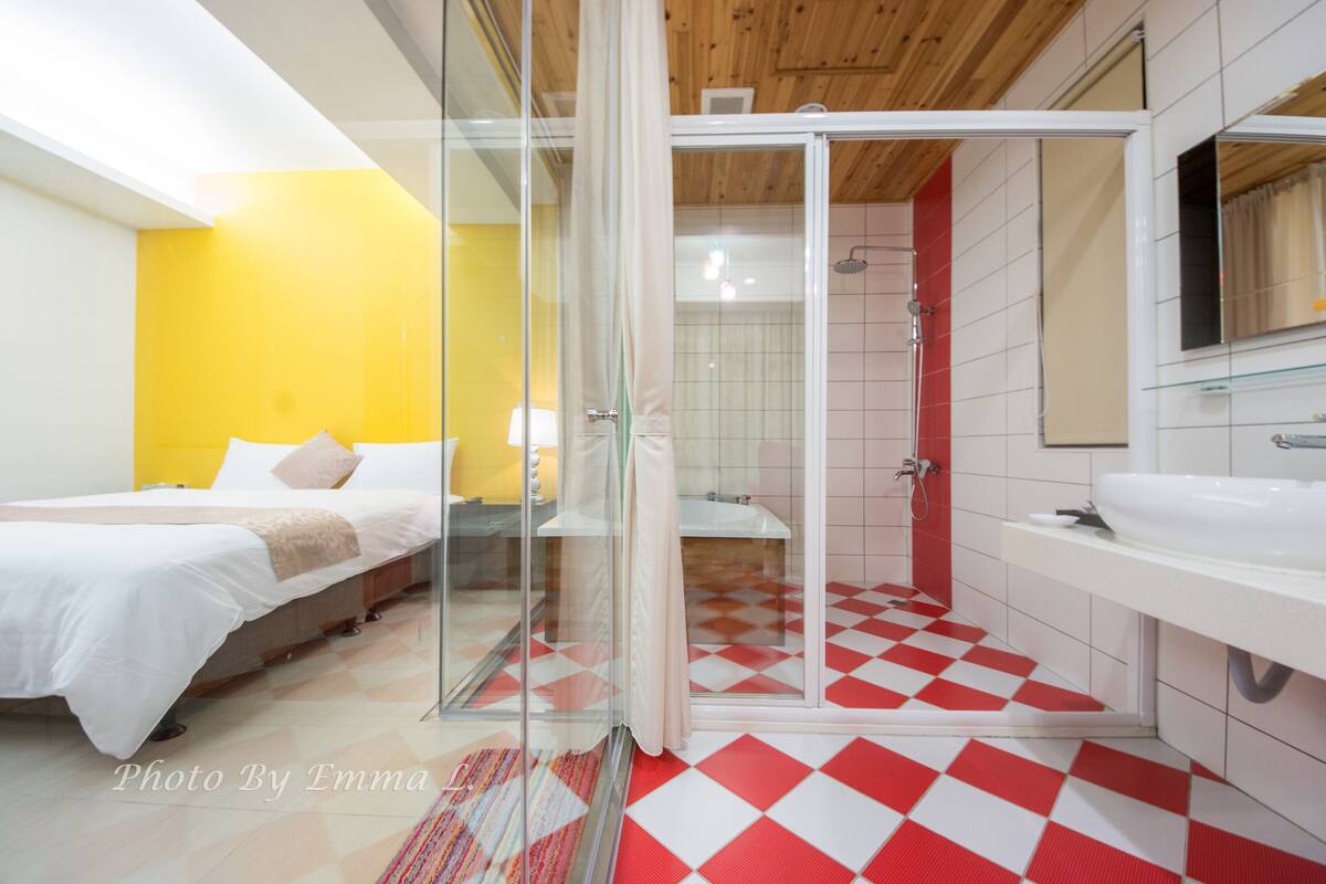 A modern hotel room with a glass shower enclosure, a bed with white bedding, a yellow accent wall, and a checkered floor.