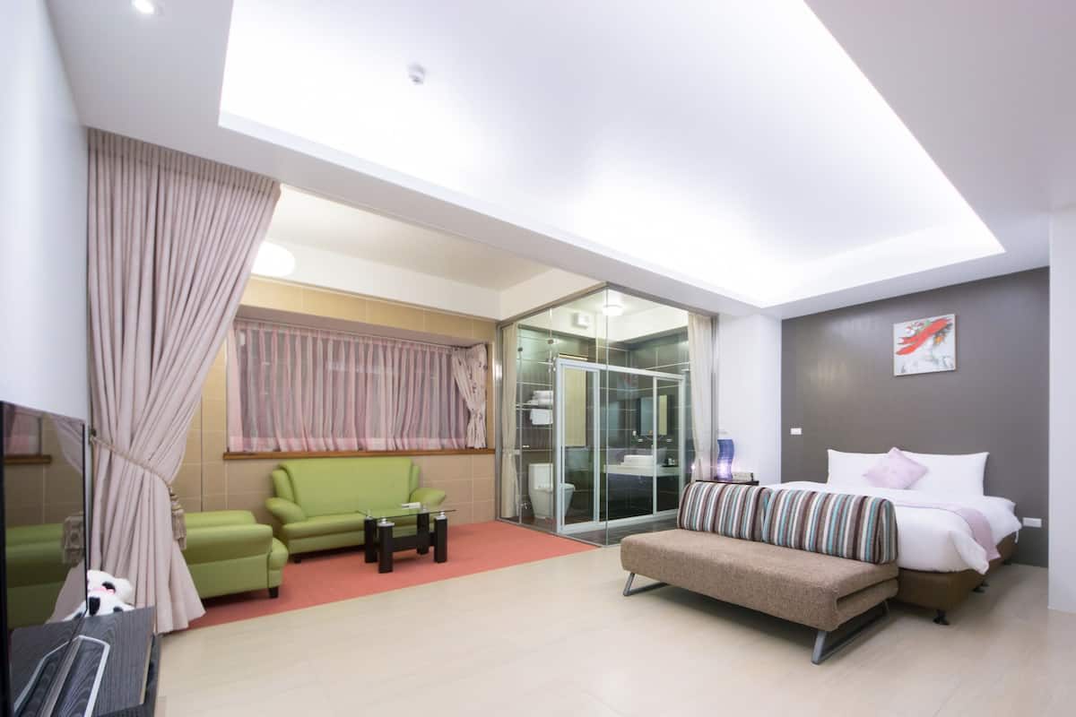 A modern hotel room with a glass shower enclosure, a green sofa, a bed with striped bedding, and a small table.