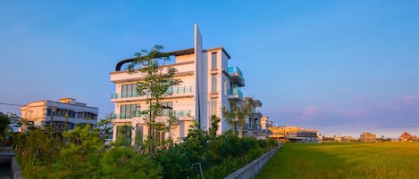 Un moderno edificio residencial de varios pisos con balcones y ventanas amplias, rodeado de vegetación y un campo de arroz.