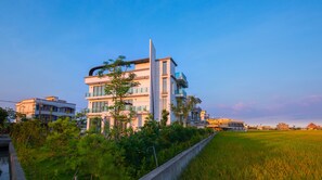 A modern multi-story residential building with balconies and large windows, surrounded by greenery and a rice field.