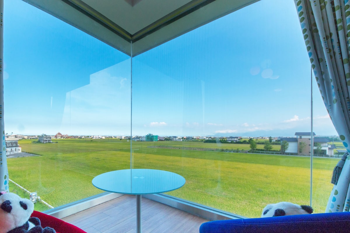 A room with a large glass window overlooking a grassy field and buildings. There is a small round table and two plush toys on a red couch.