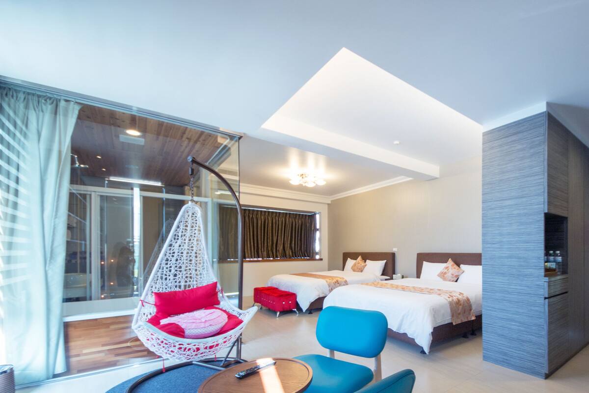 A modern hotel room with a large bed, a hanging chair, a blue armchair, a small table, and a large window with curtains.