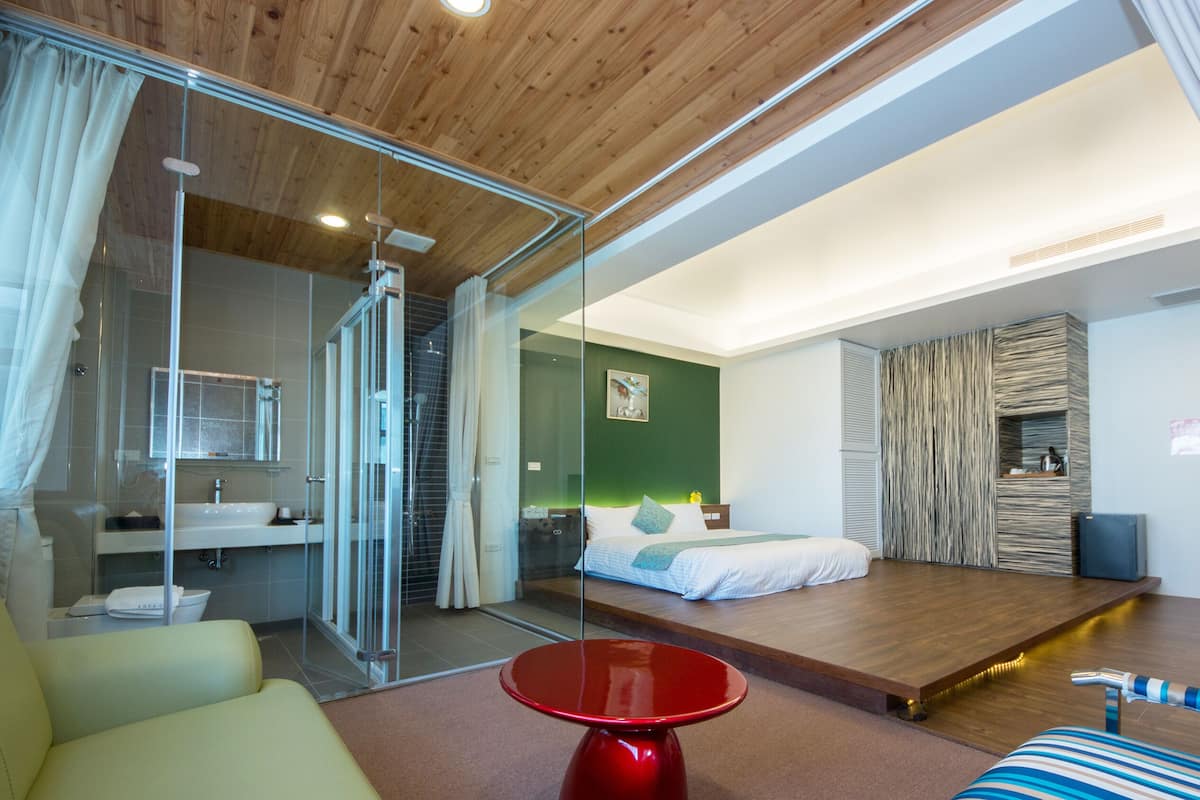 A modern hotel room with a glass shower enclosure, a bed with a wooden headboard, a red table, and a striped sofa.