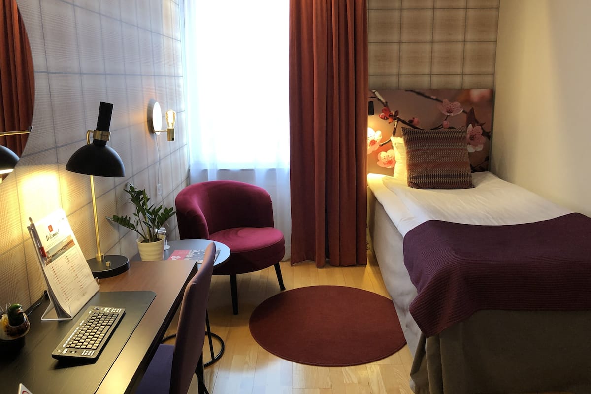 Quarto de hotel com secretária, cadeira, computador, candeeiro e cama com cobertor.