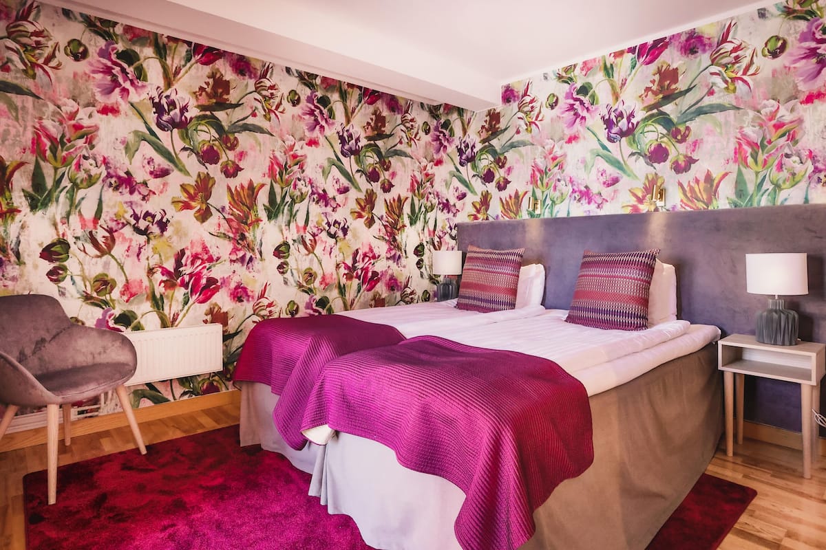 Um quarto com papel de parede floral, uma cama com manta roxa, uma cadeira cinza e uma pequena mesa de cabeceira com um abajur.