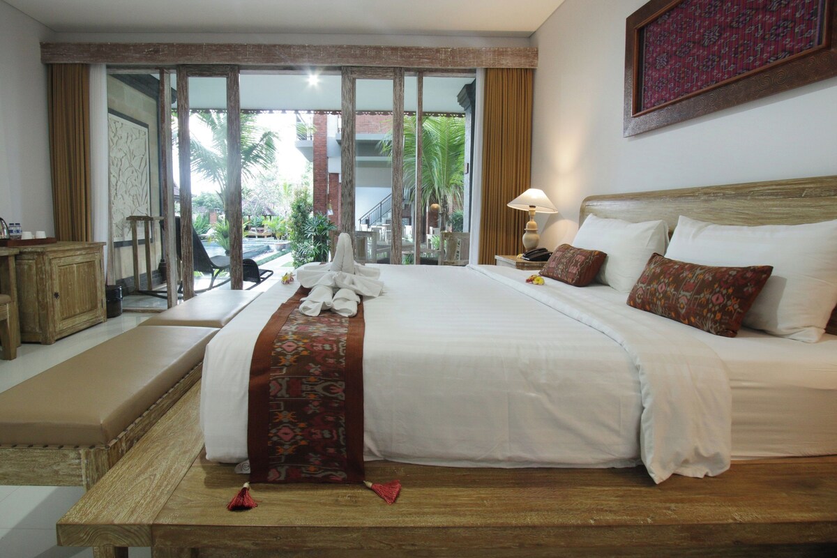 A hotel room with a large bed, a wooden bench, a desk, and a view of the outdoors.