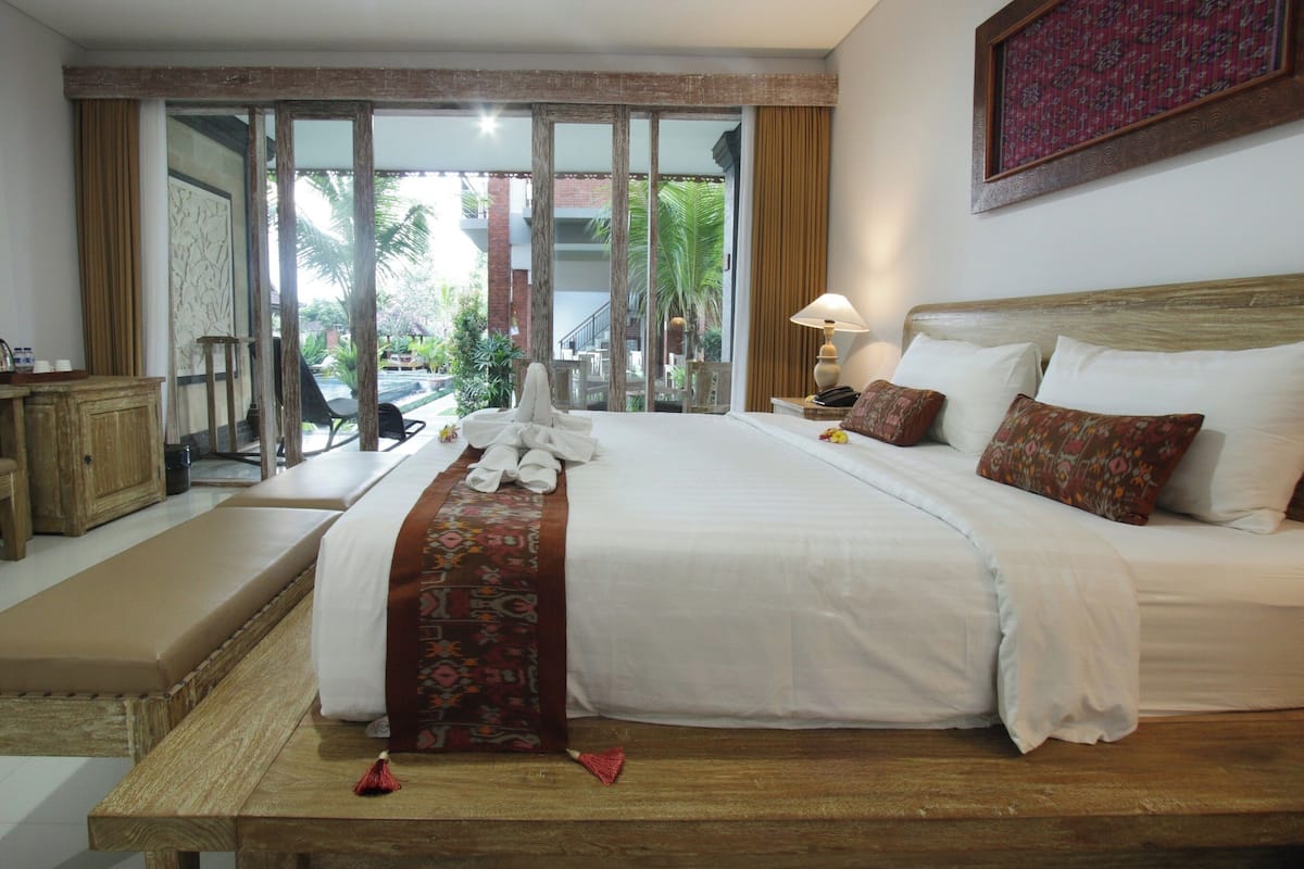 A hotel room with a large bed, a wooden bench, a desk, and a view of the outdoors.