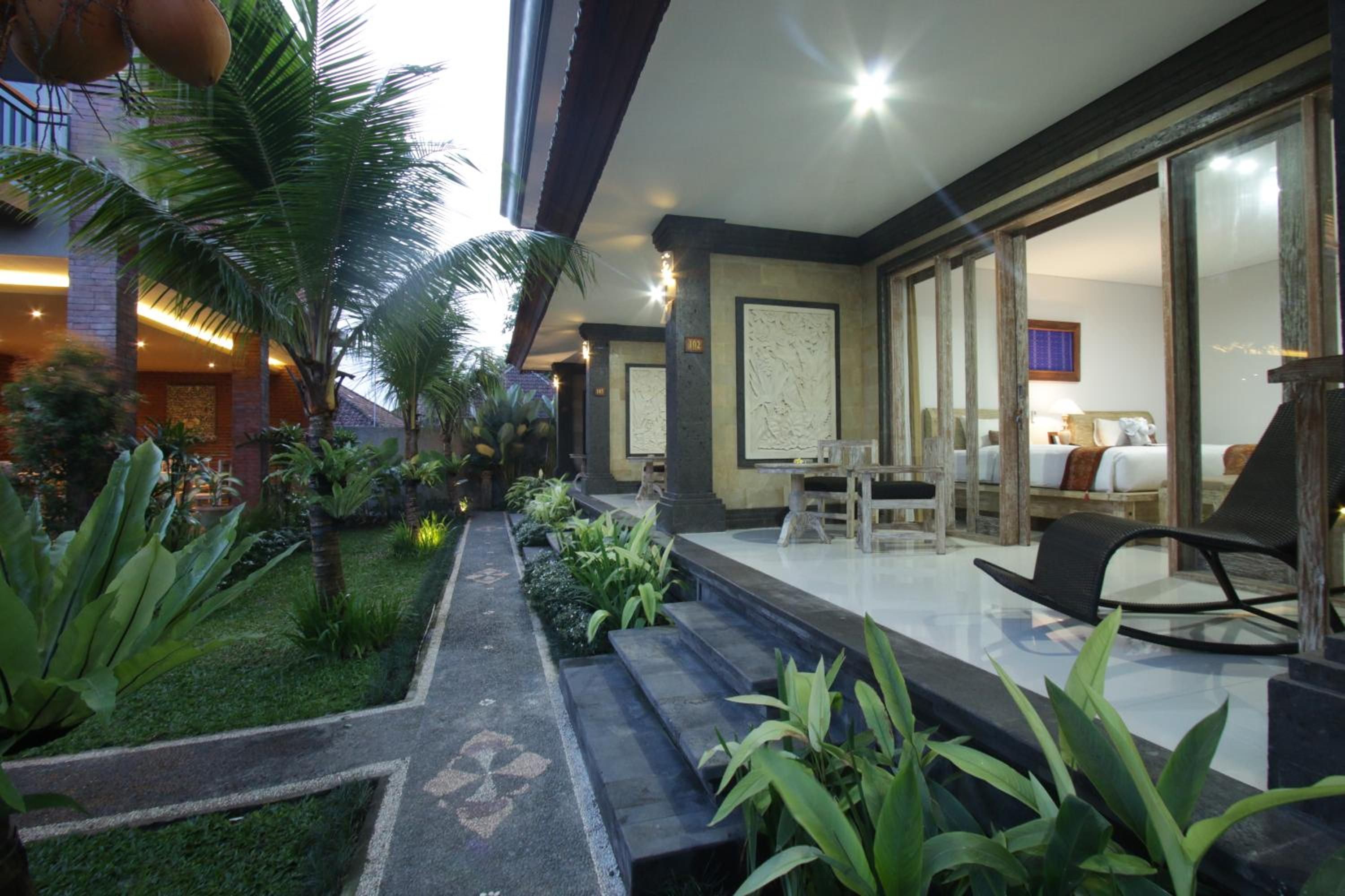 A hotel corridor with a glass door leading to a room, a small outdoor seating area with a table and chairs, and a well-maintained garden with palm trees and plants.
