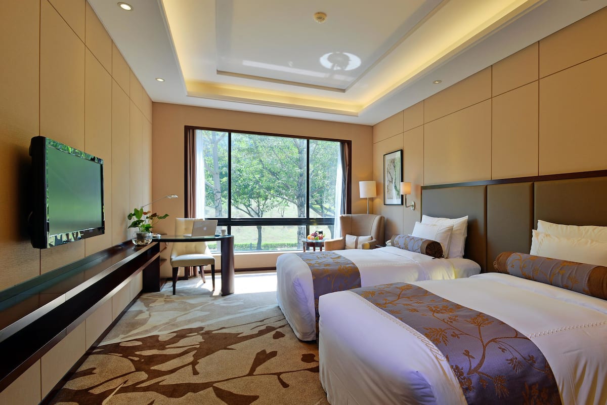 A hotel room with two beds, a desk, a TV, and a large window with a view of trees.