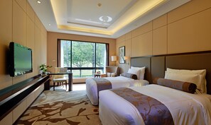 A hotel room with two beds, a desk, a TV, and a large window with a view of trees.
