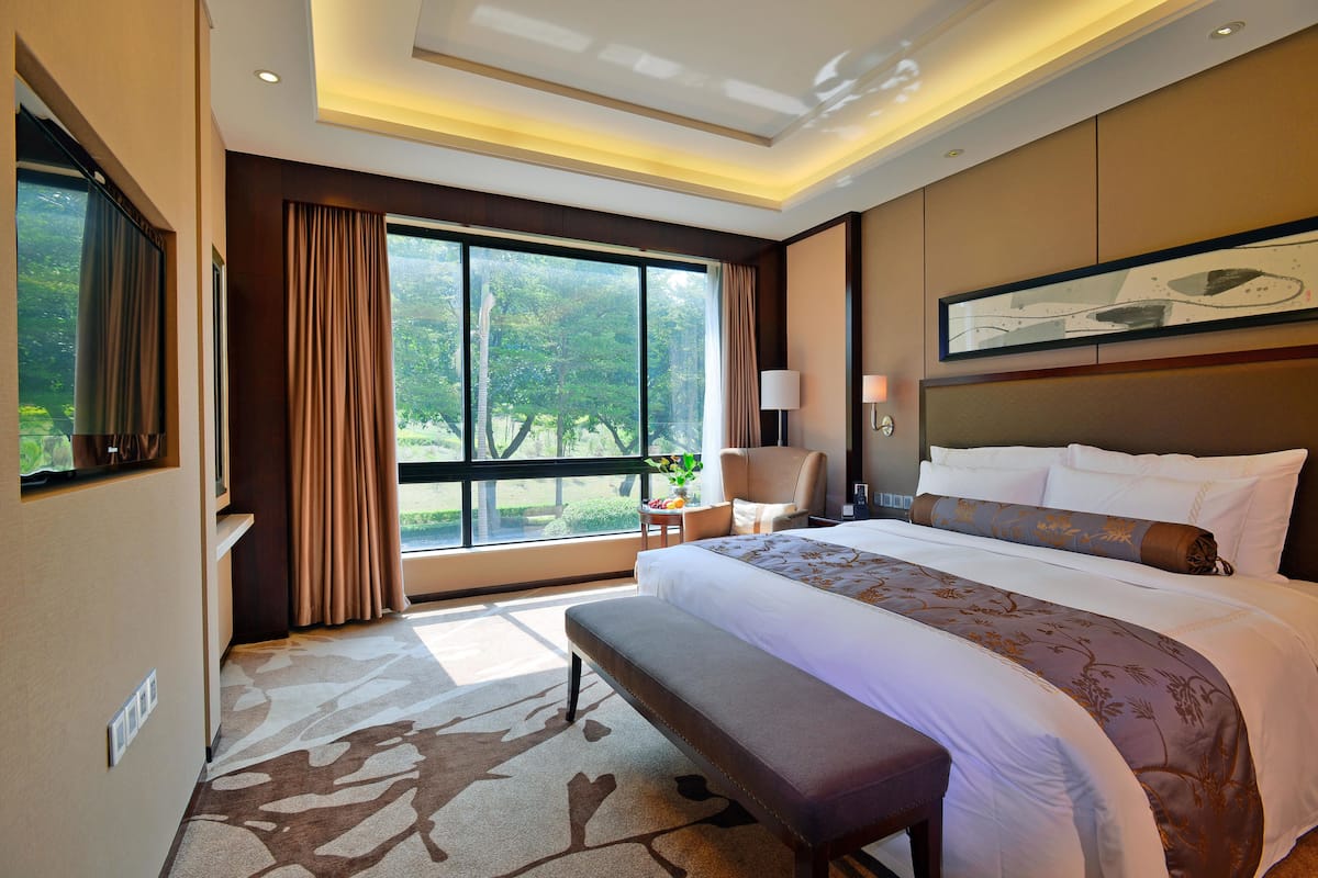 A hotel room with a large bed, a bench, a desk, and a view of greenery outside.