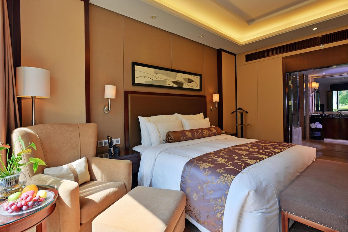 A hotel room with a large bed, a sofa, a small table with fruit, and a view of the outdoors.