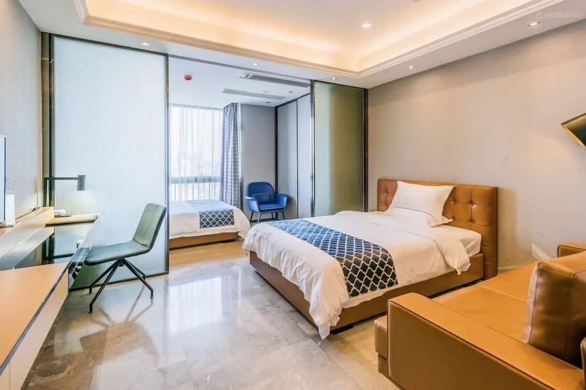 A modern hotel room with a large bed, a desk, a chair, and a glass partition.