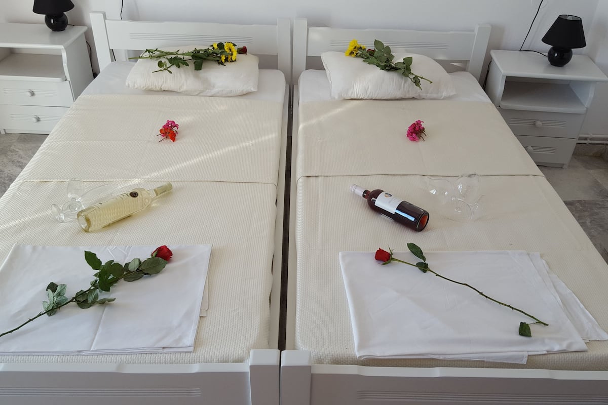 A hotel room with two beds, each with white linens and a single red rose. There are bedside tables with lamps, a small white nightstand, and a white dresser.