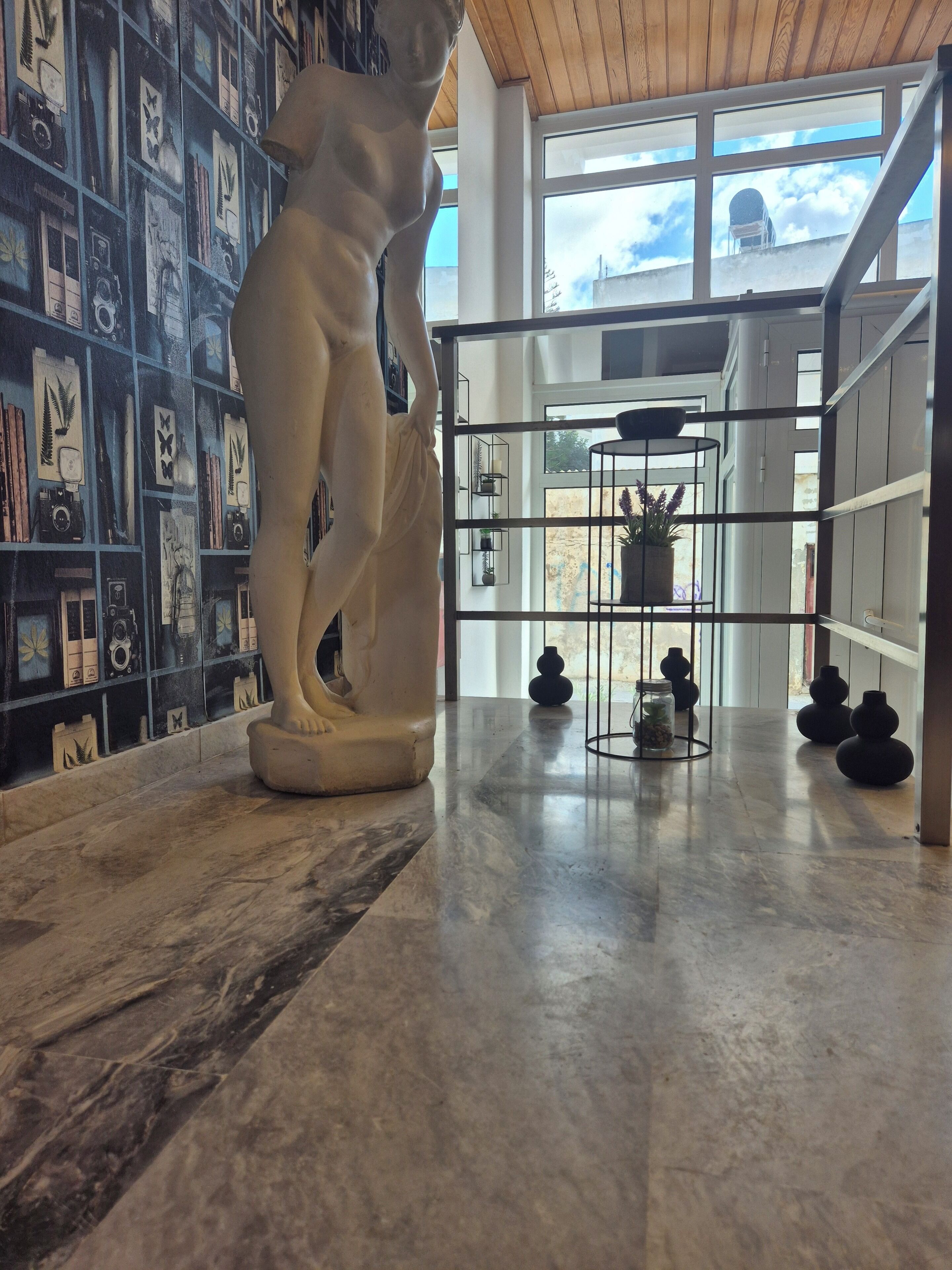 A marble floor with a white statue, a wall with framed pictures, and a view of the sky through a window.