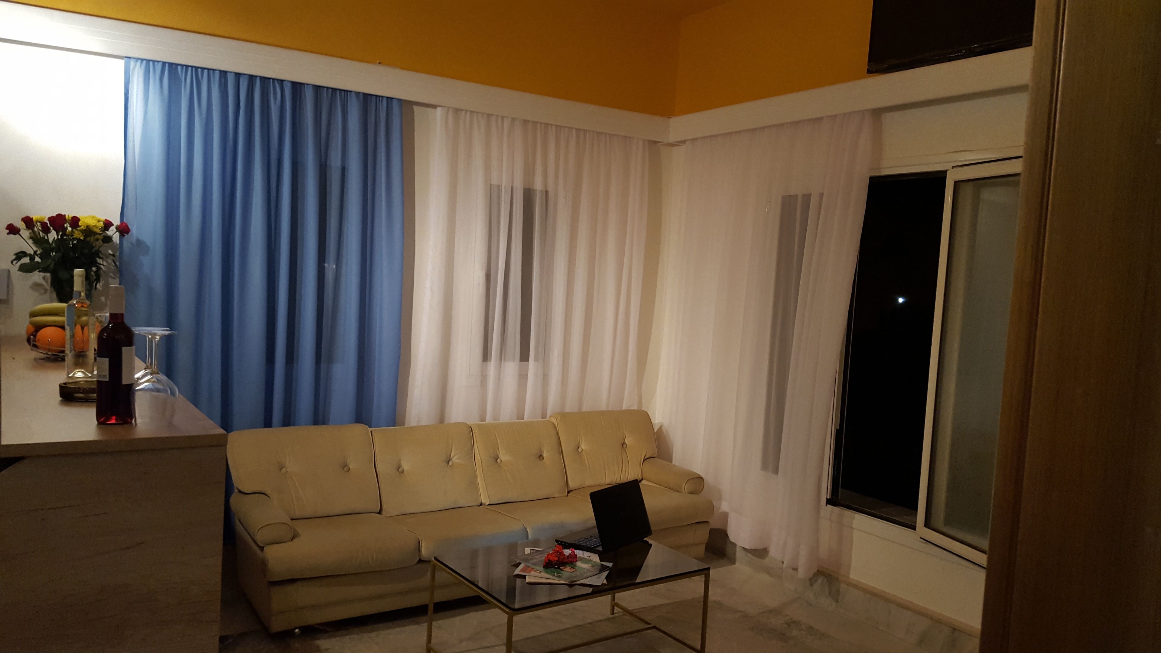 Una sala de estar con un sofá color beige, una mesa de centro con una computadora portátil y una ventana con cortinas.