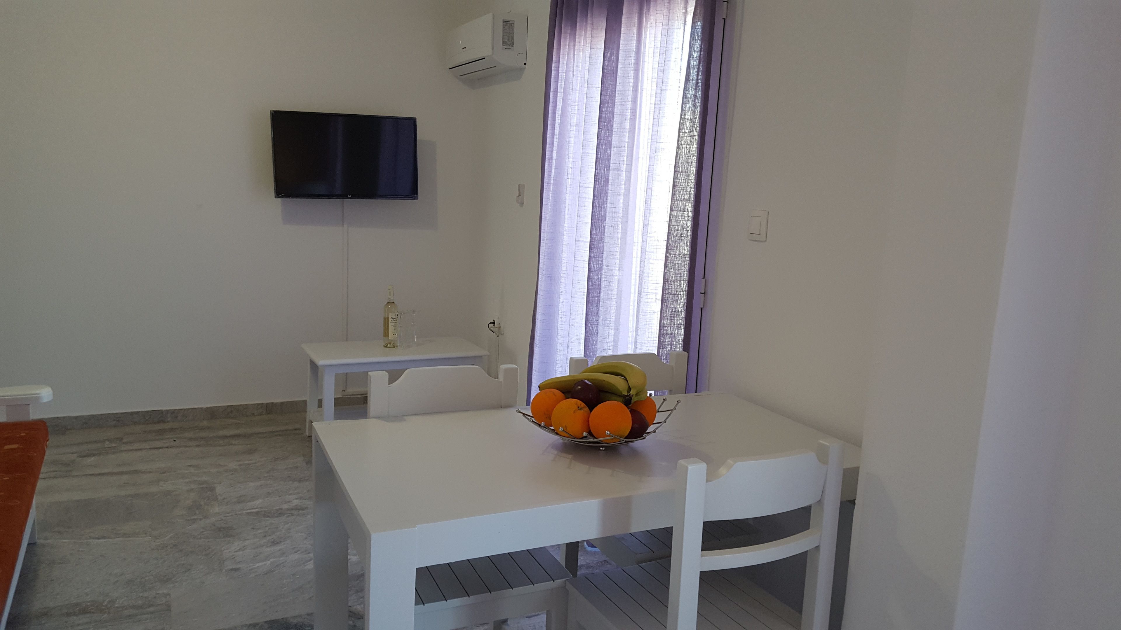 A small, clean room with a white table, a white chair, a flat-screen TV mounted on the wall, and a window with sheer curtains.