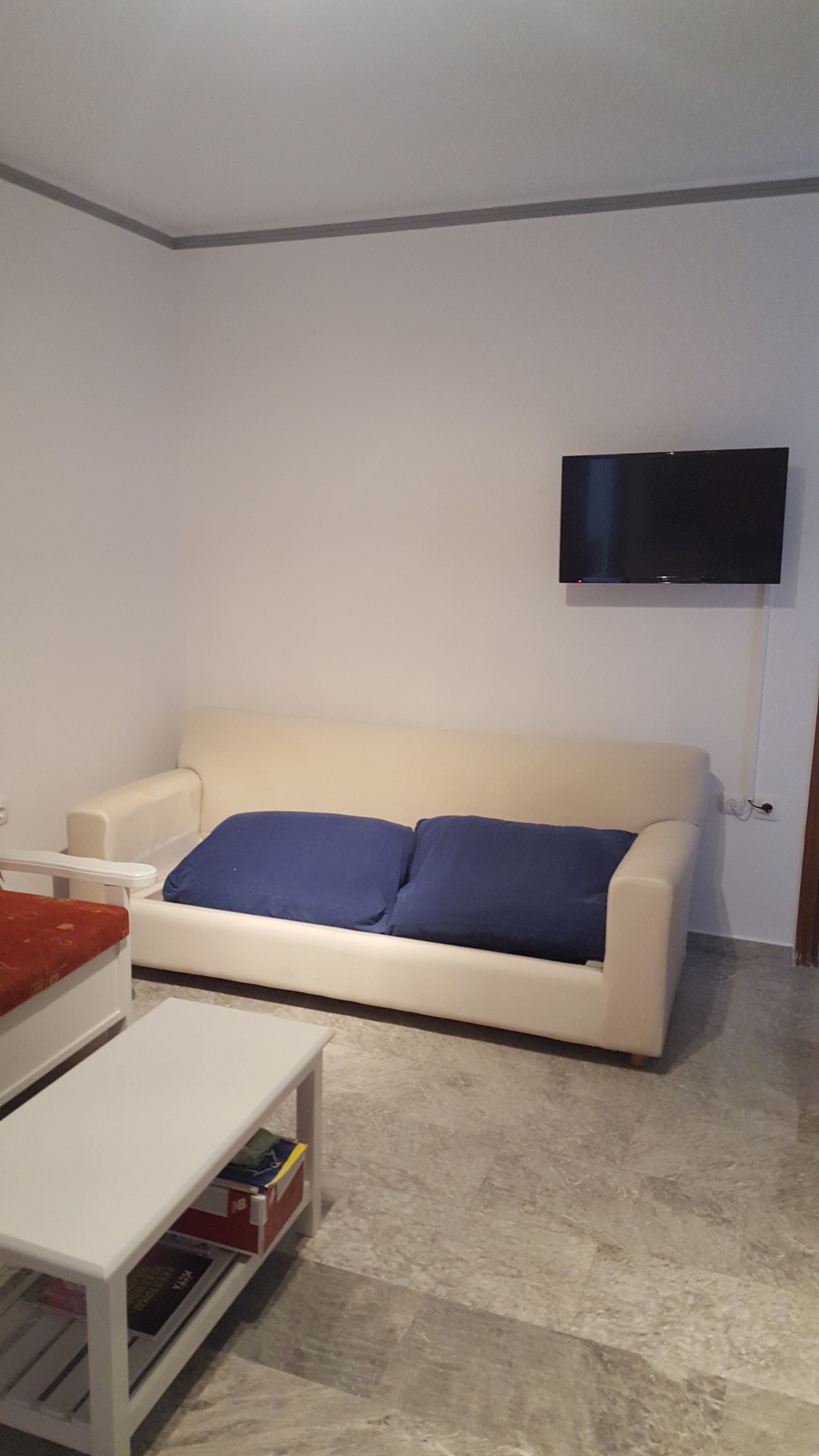 A living room with a white sofa, a flat-screen TV mounted on the wall, and a small white table with a red cushion.