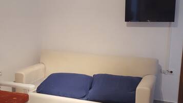 A living room with a white sofa, a flat-screen TV mounted on the wall, and a small white table with a red cushion.