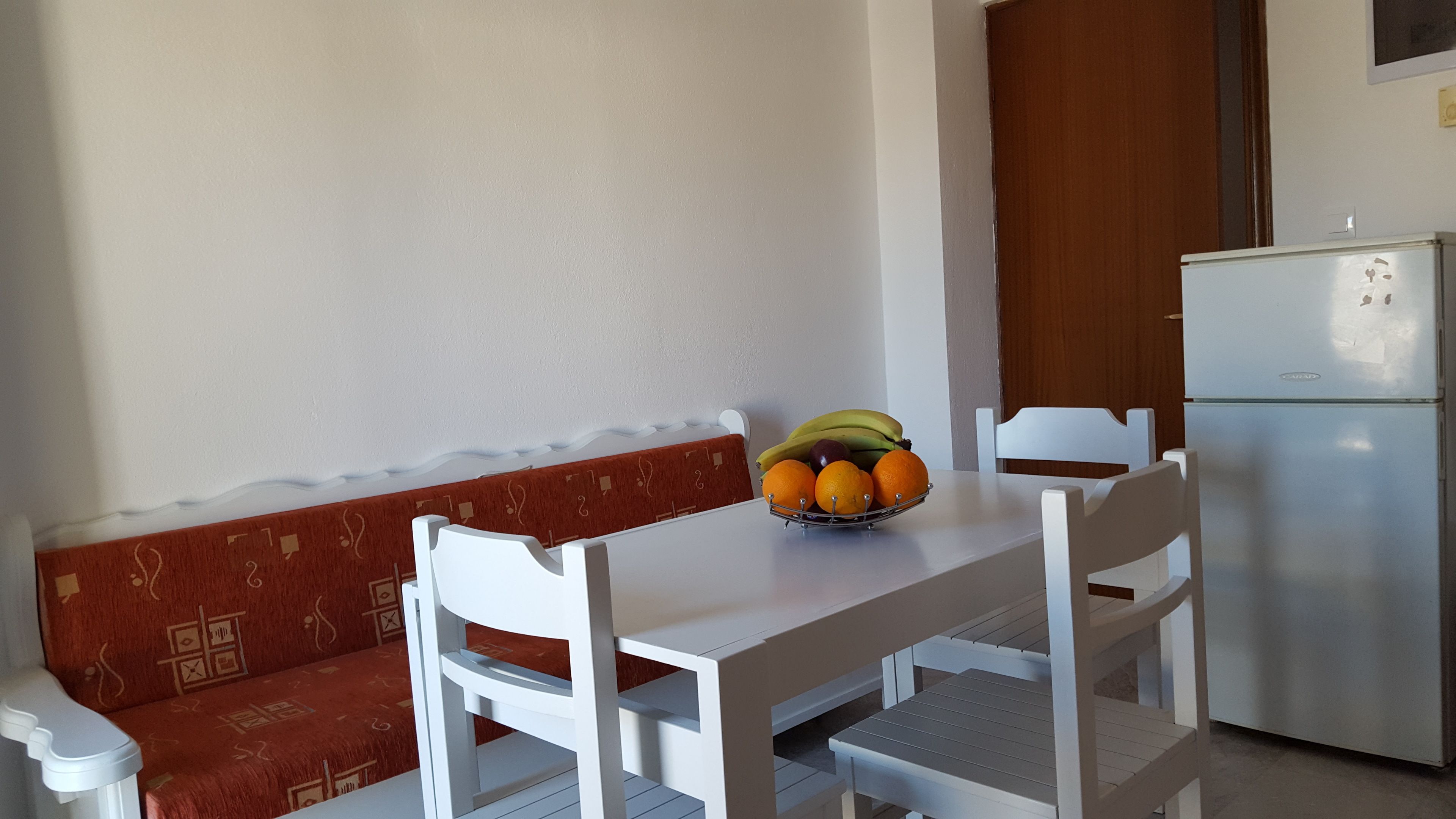 Une petite cuisine équipée avec une table blanche, deux chaises blanches, un petit panier de fruits et un réfrigérateur.