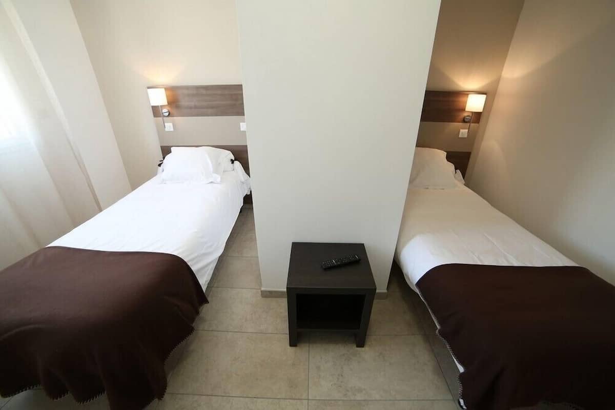 Quarto de hotel com duas camas de solteiro, cada uma com colcha marrom e travesseiros brancos. Há uma pequena mesa de cabeceira preta entre as camas, com um controle remoto sobre ela.