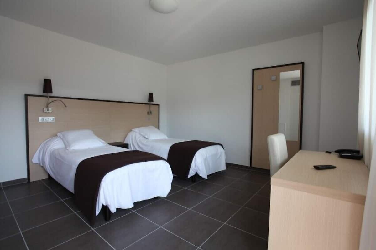 Quarto de hotel com duas camas de solteiro, uma escrivaninha, uma cadeira e um espelho.
