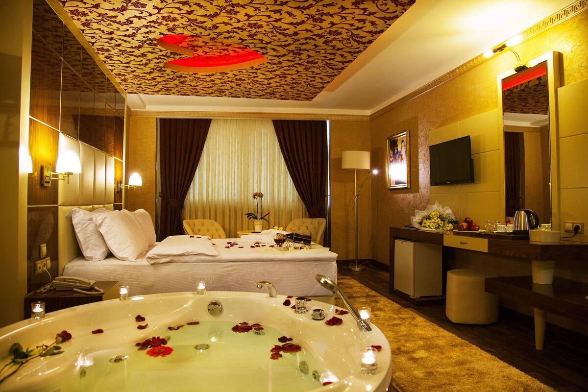 Habitació d'hotel amb un llit gran, una zona de seients amb un sofà i butaques, una taula petita amb un vas de flors, una televisió i un bany amb banyera i mirall.