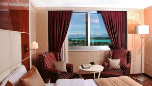 A hotel room with a bed, two armchairs, a small table with a lamp, and a view of a body of water and mountains. - Cukurova Erten Otel (Adana)