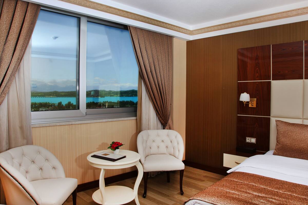 Habitació d'hotel amb una finestra gran, dues cadires, una taula petita amb una flor, un llit i una nitòria.