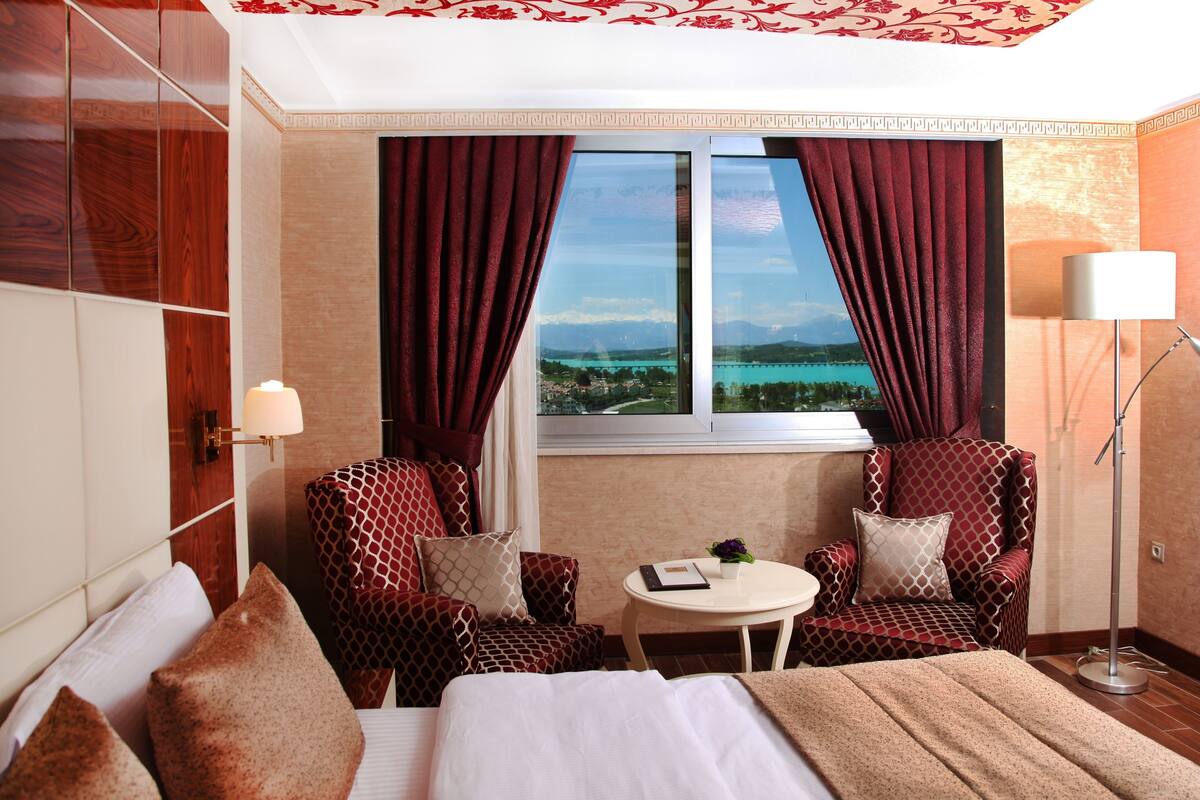 Habitació d'hotel amb un llit, dues butaques, una taula petita, una làmpada i una finestra que mostra un cos d'aigua i muntanyes.