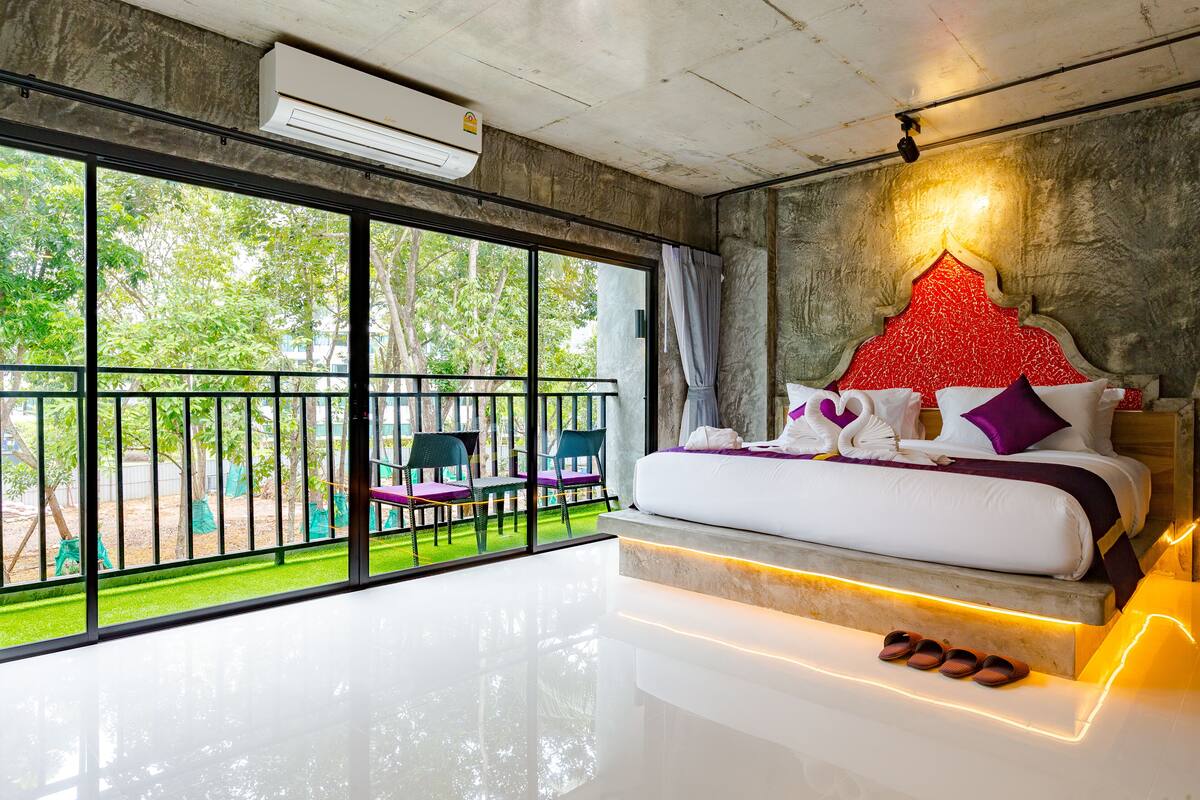 A modern bedroom with a large window, a balcony with chairs, and a bed with pillows and a blanket.
