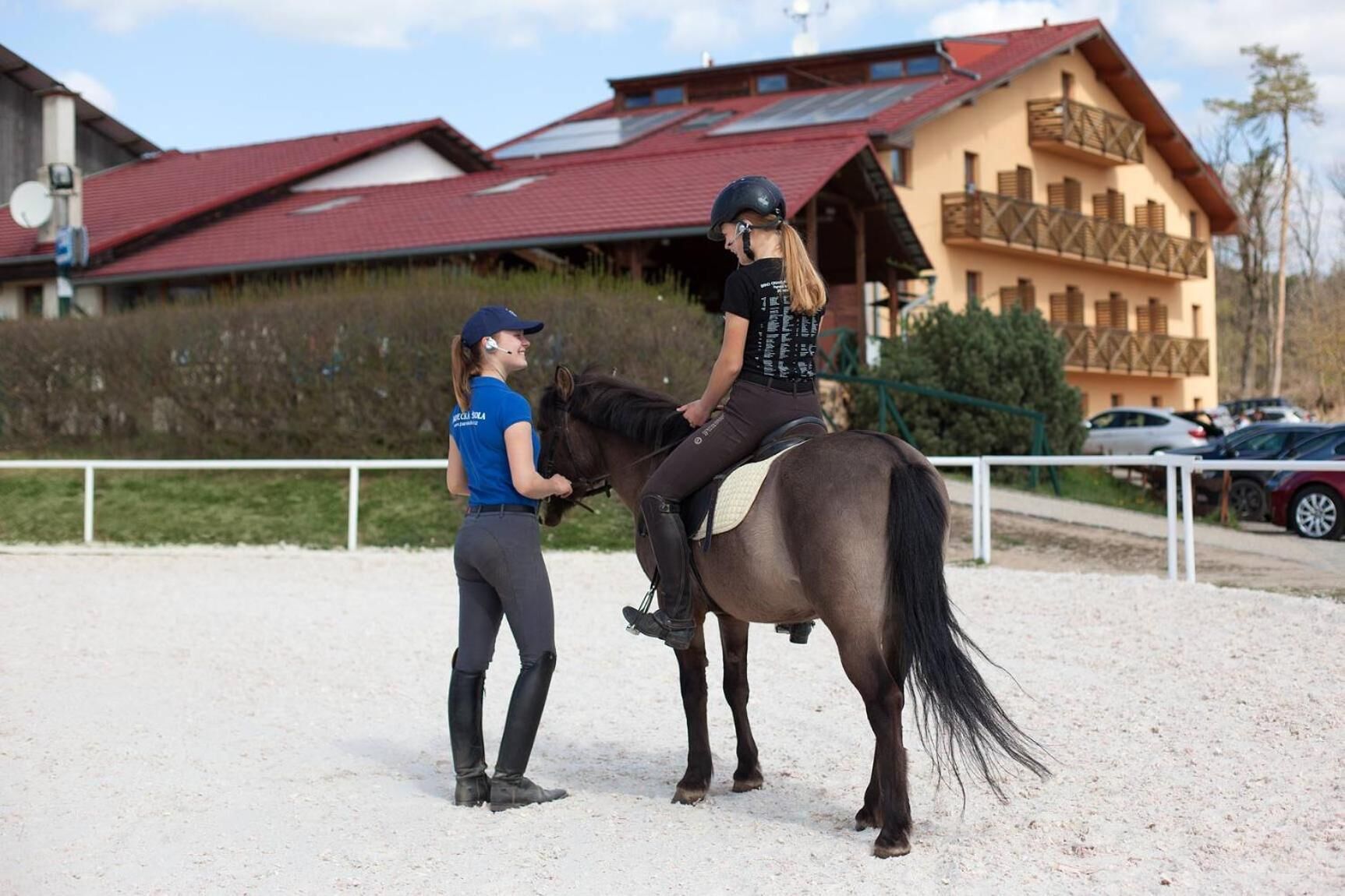 En person rider på hest i en udendørs arena med bygninger i baggrunden.