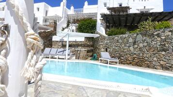 Un área de piscina con suelo de piedra, edificios blancos al fondo y una columna blanca con una cuerda.
