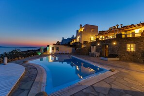 A pool area with a stone deck, a clear pool, and a view of the sea at dusk. - SeaBlue Villas (Mykonos)