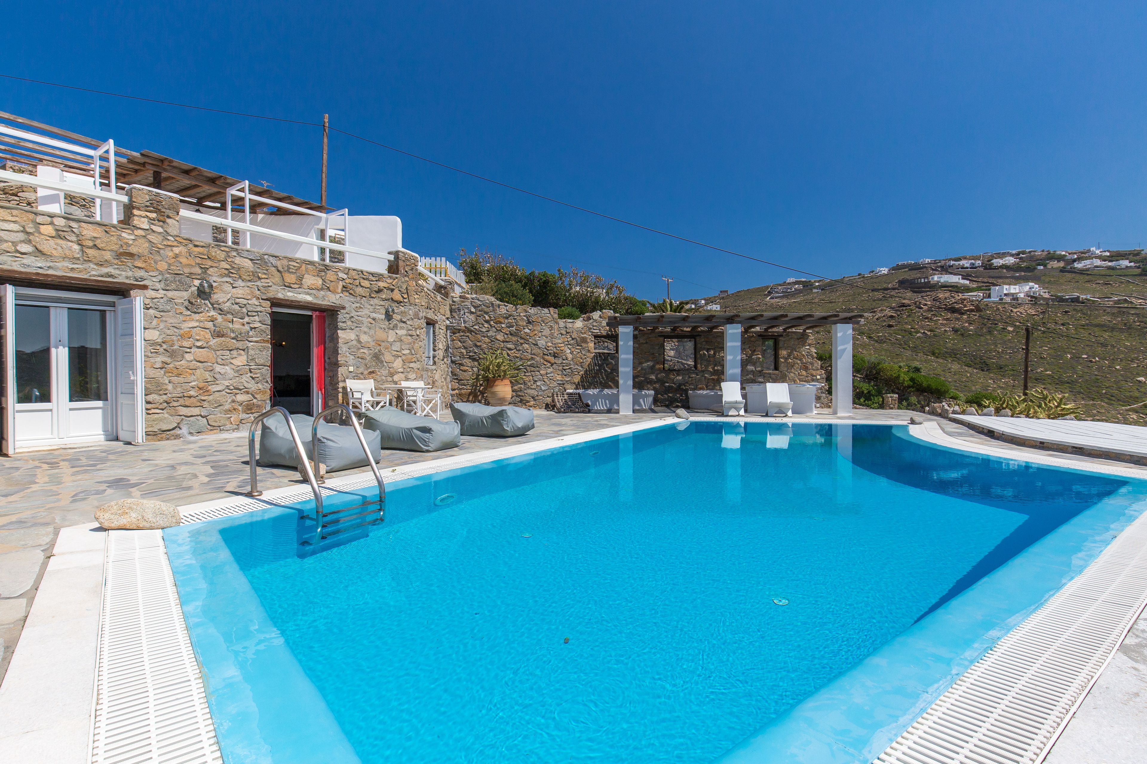 Una piscina con revestimiento de piedra, una zona de estar cubierta y vistas a una colina.