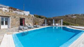 Una piscina con revestimiento de piedra, una zona de estar cubierta y vistas a una colina.