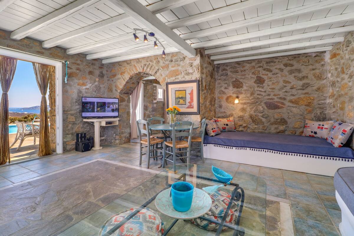 Un espacio de sala acogedor con paredes de piedra, una mesa de vidrio con una taza azul, un sofá con cojines estampados y un televisor en una pared de piedra.