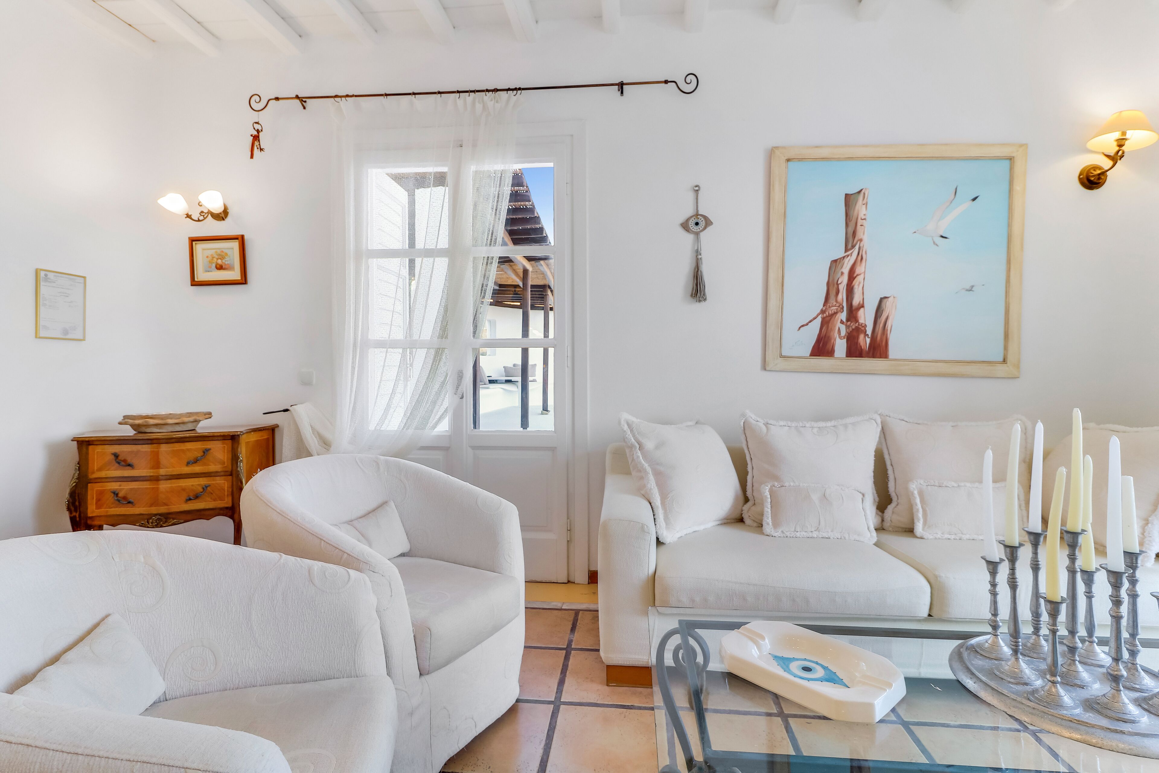 A living room with white furniture, a wooden side table, a glass coffee table, a wall-mounted lamp, and a framed picture of trees and birds.