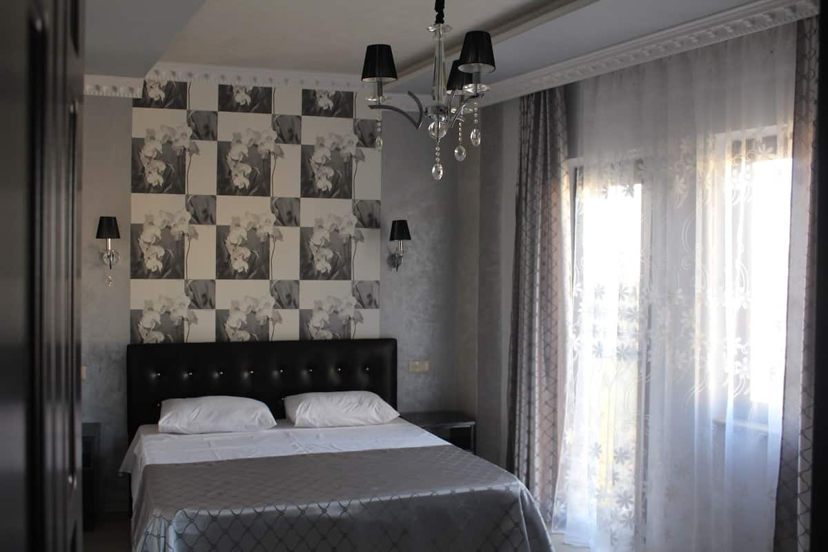 A bedroom with a checkered floral wallpaper, a large bed with white linens, a chandelier, and sheer curtains.