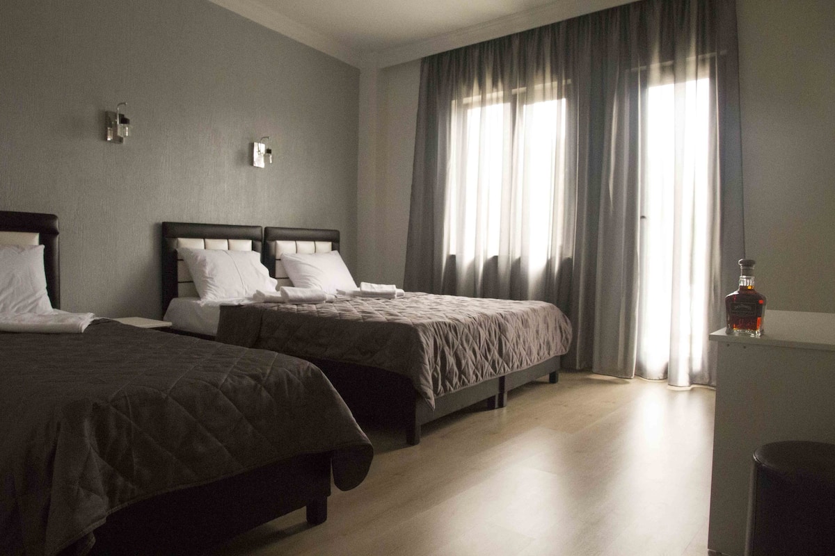A hotel room with two beds, a large window with curtains, a small table with a bottle, and a trash bin.