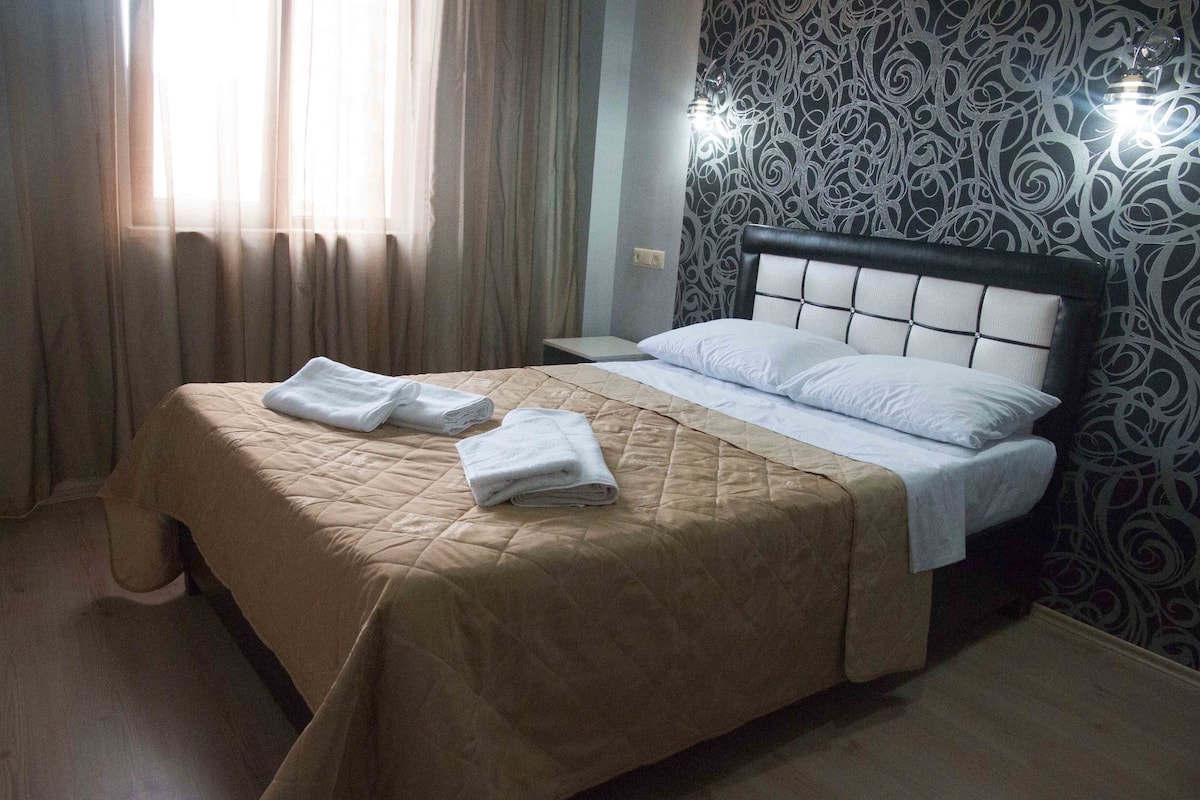 A neatly made bed with a beige bedspread and two folded white towels, a bedside table, and a wall with a decorative pattern.