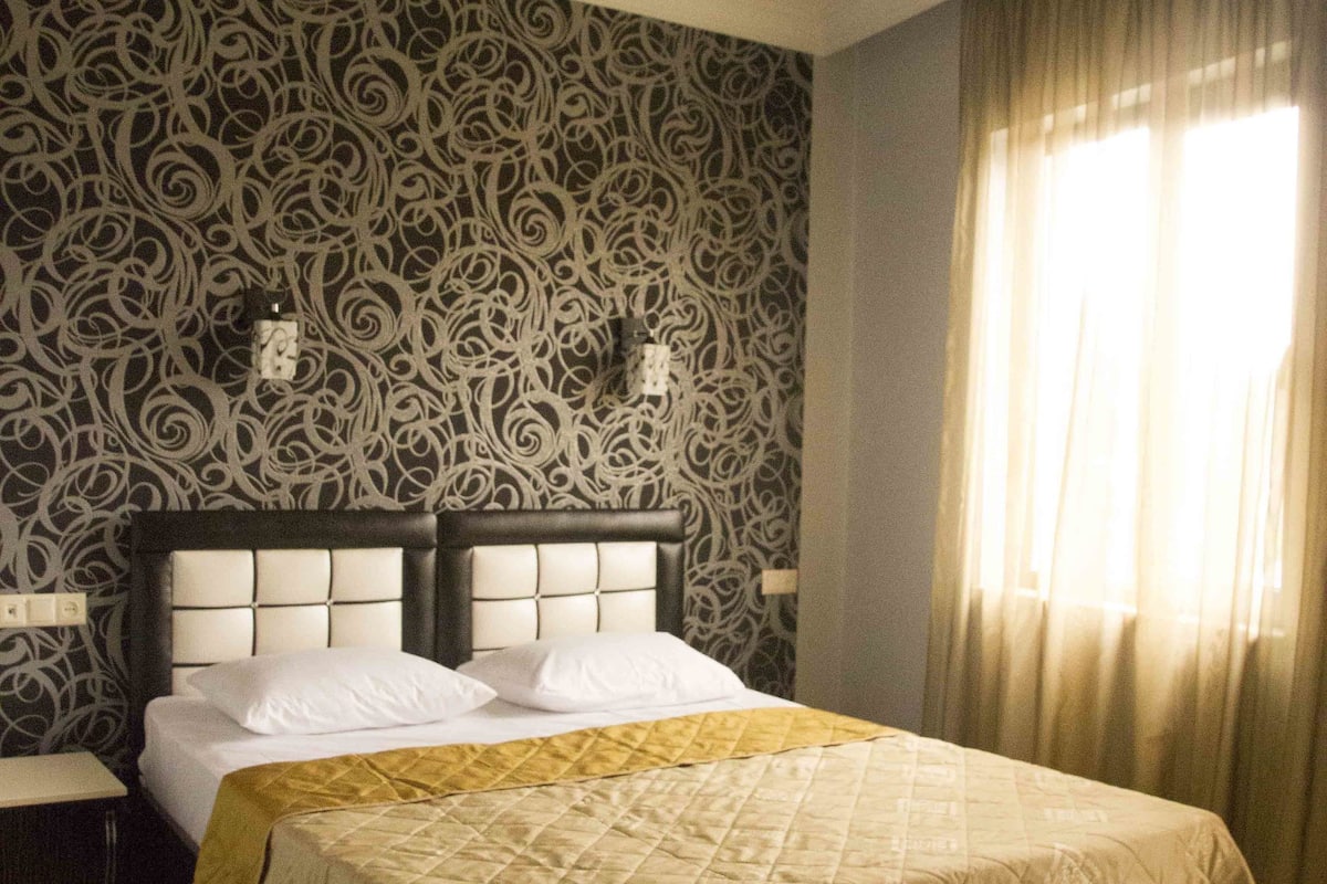 A bedroom with a large bed, a nightstand, patterned wallpaper, and a window with curtains.
