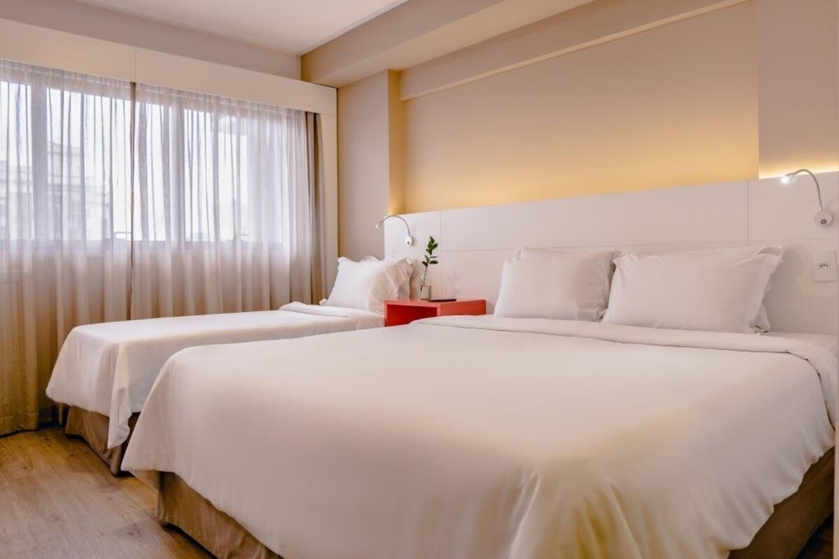 Habitación de hotel con dos camas, un ventanal con cortinas traslúcidas, una mesita de noche con lámpara y una lámpara de pared.