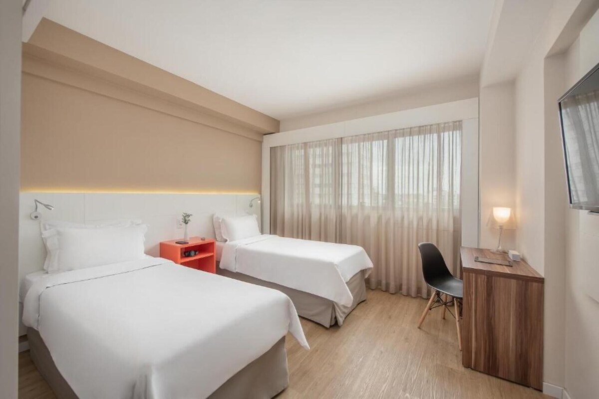 Habitación de hotel con dos camas, un escritorio, una silla, una mesita de noche y un ventanal con cortinas.