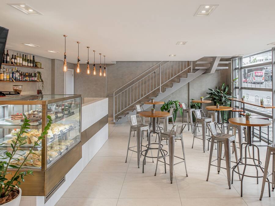 Um café moderno com uma vitrine de doces, banquetas de metal e uma escada que leva a um andar superior.