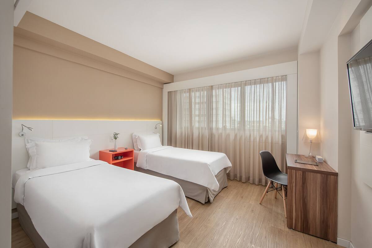 Een hotelkamer met twee bedden, een bureau, een stoel en een raam met gordijnen.