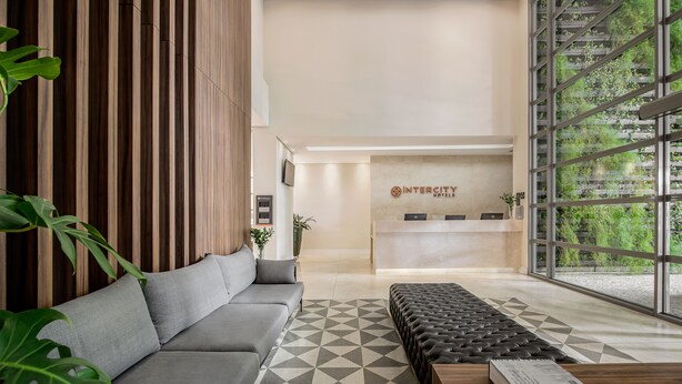 Een moderne hotellobby met een receptiebalie, een grijze bank, een kamerbreed tapijt met dessin en een groot raam met uitzicht op groen.